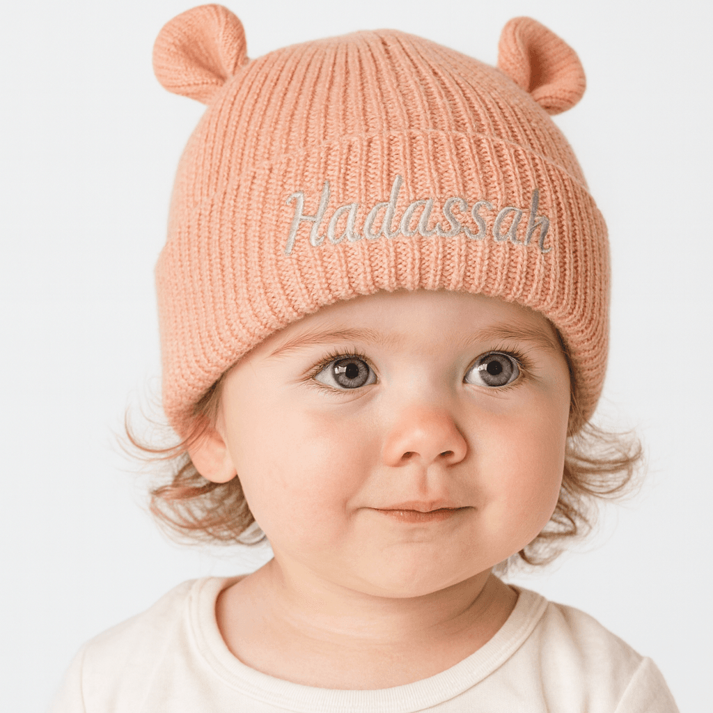 Gebreide baby muts in de kleur perzik met schattige oortjes en de naam Hadassah geborduurd op de omslag.
